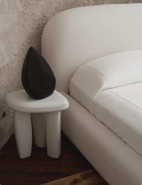Pantheone Obsidian black wireless speaker on modern white nightstand beside curved bed in soft-toned bedroom