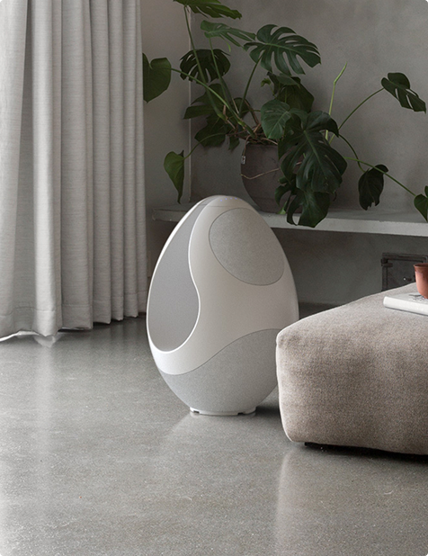 Pantheone I white wireless speaker placed on polished concrete floor beside ottoman and indoor plant in modern living room