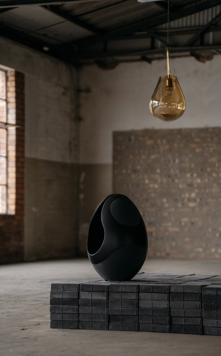 Black Pantheone I speaker on a concrete surface with a hanging gold light bulb in an industrial setting.