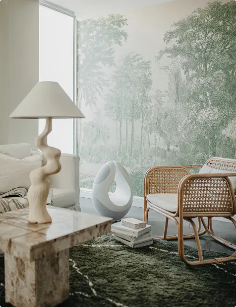 A styled living room corner featuring a large wall mural of a misty forest, a Pantheone I wireless speaker in white, a table lamp on a stone-topped table, two woven rattan chairs, a green shag rug, and a white abstract sculpture.