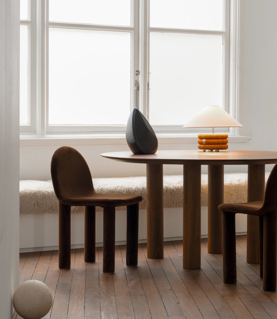 Wooden dining table with chairs in a bright room with the Obsidian wireless speaker in black and large windows.
