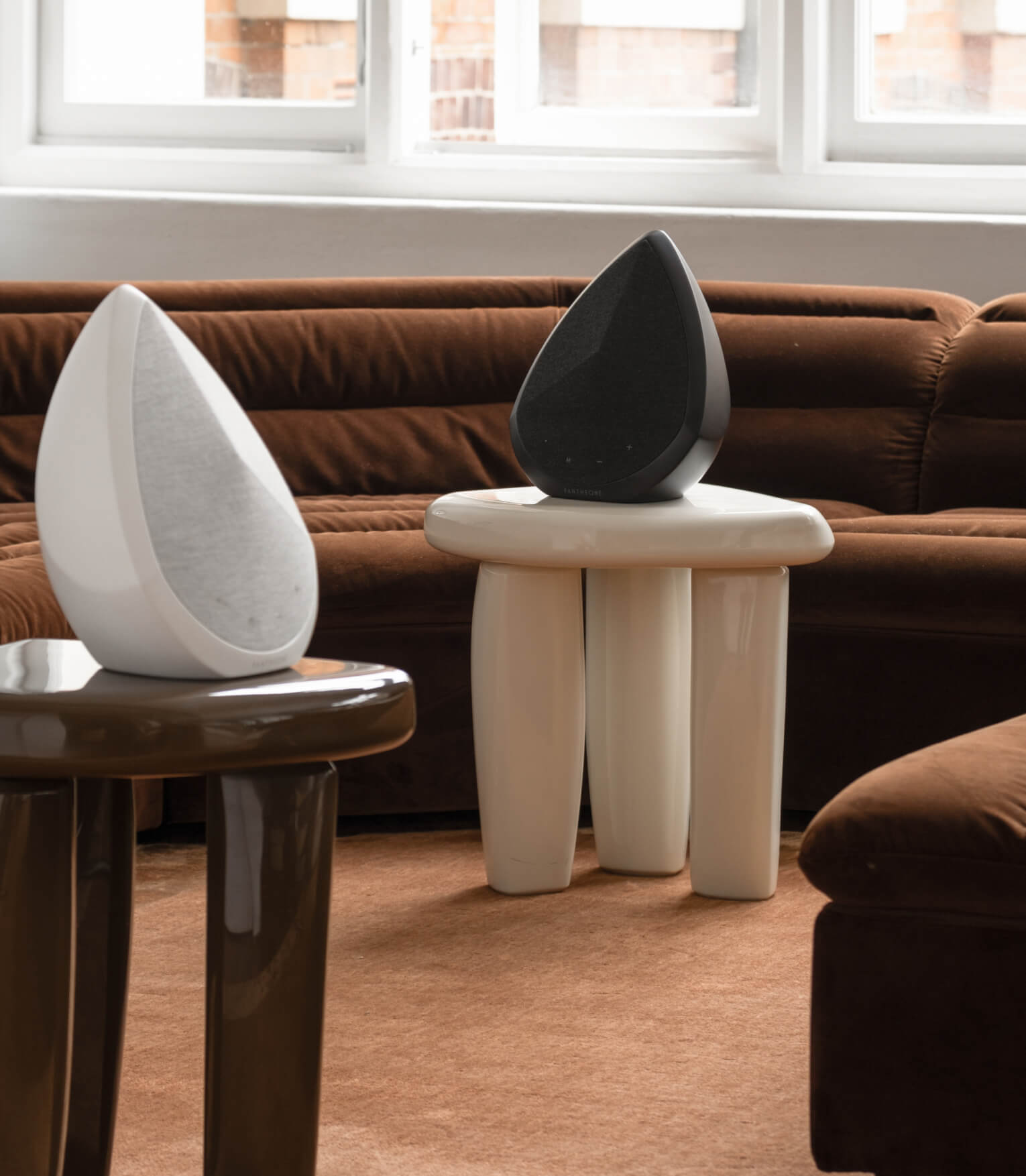 White and black Pantheone Obsidian wireless speakers styled on sculptural side tables in a contemporary living room with curved velvet sofa and natural light