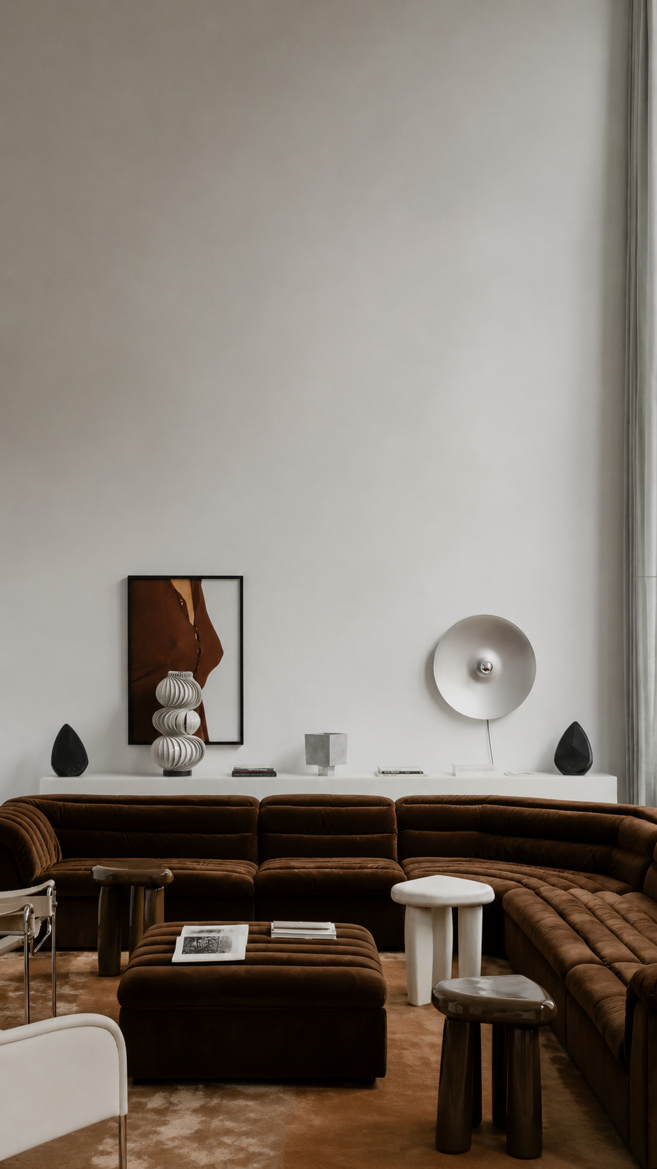 A modern living room featuring the Obsidian wireless speakers in a stereo pair mode.With a sofa with matching ottomans, styled with white side tables, sculptural wall lamps, and a framed abstract print on a light grey wall.