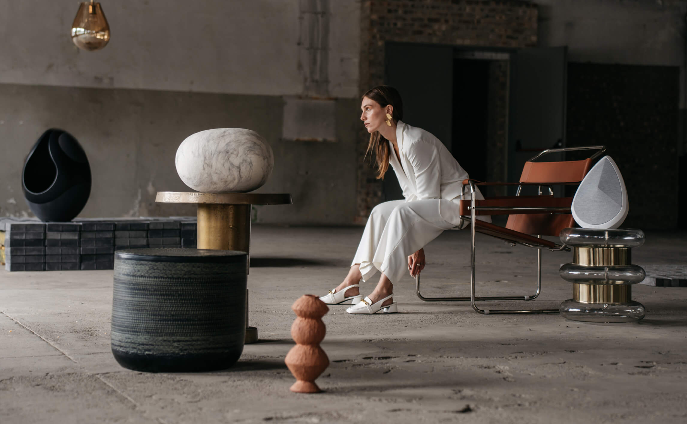 Woman in a white outfit seated in a modern industrial space, surrounded by sculptural furniture and two Pantheone speakers in black and white