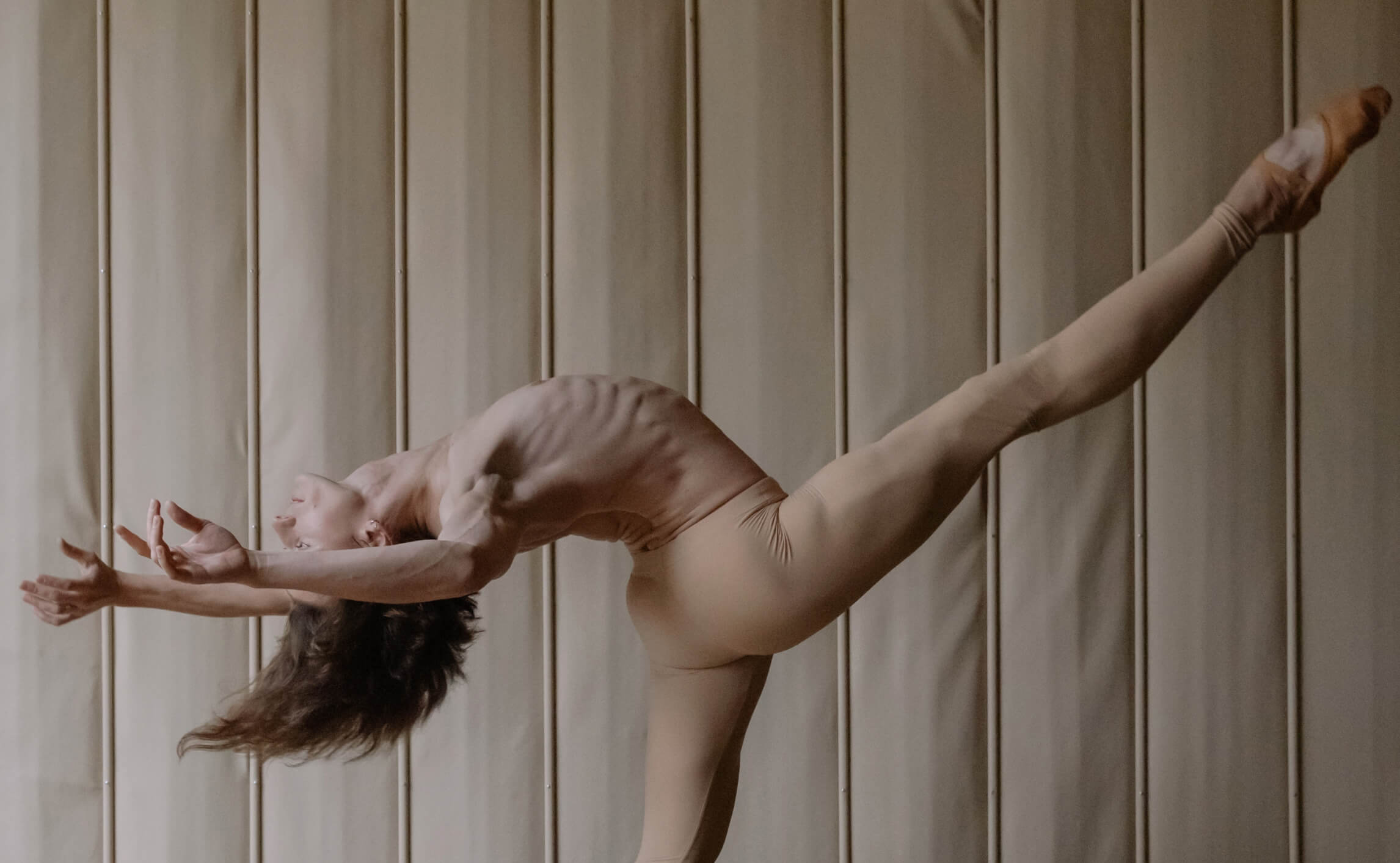 Ballet dancer performing an elegant arabesque pose against a softly lit padded wall.