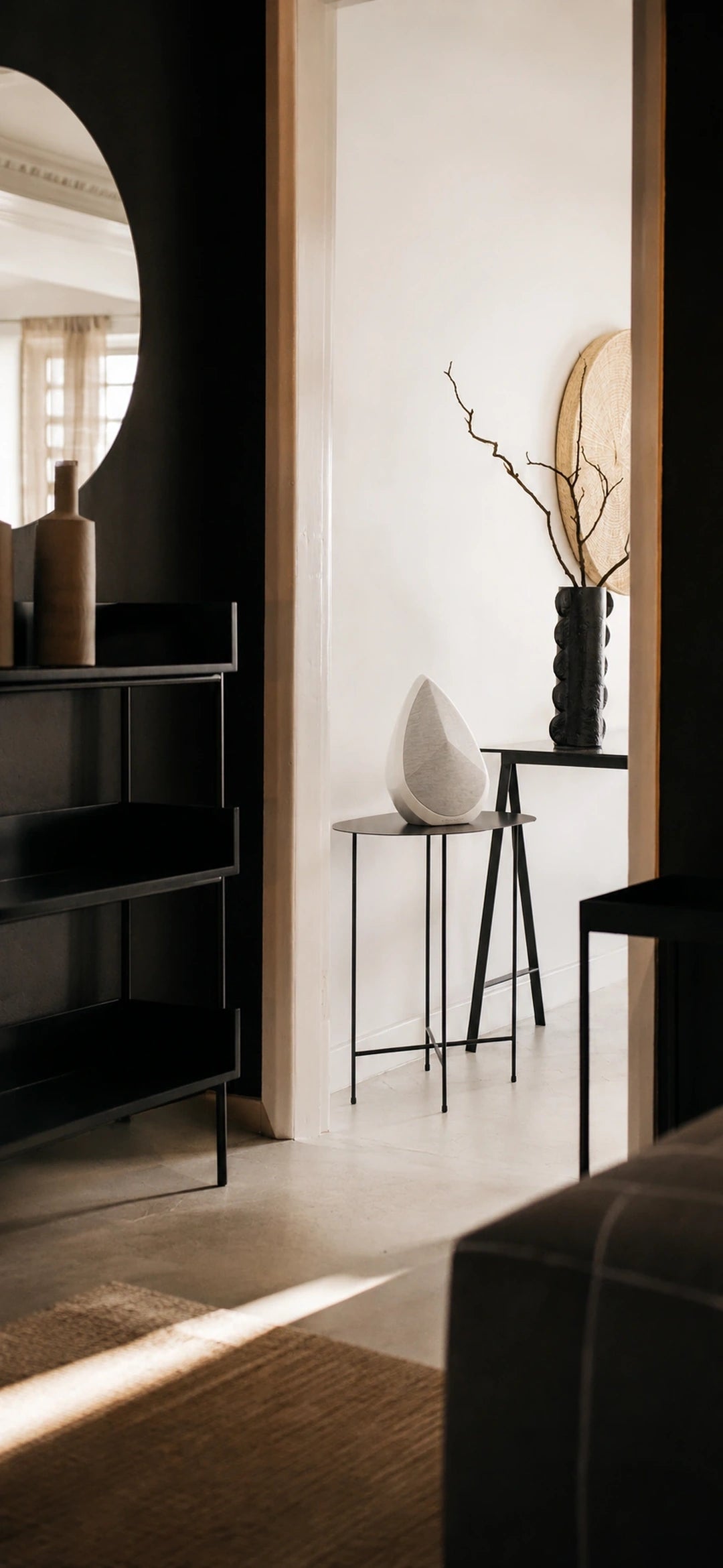 A modern interior scene with dark wood furniture, a black shelving unit, a small black side table with the White Obsidian speaker, and a tall black vase with dried branches. A large oval mirror is visible on the left, and warm sunlight streams across a light floor.