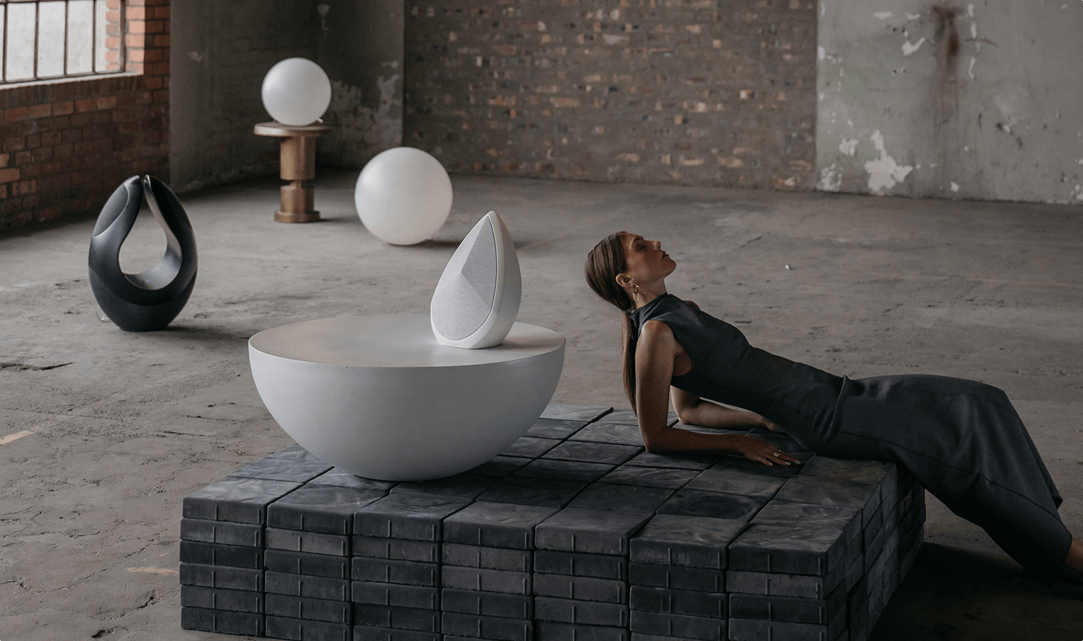 obsidian white wireless speaker on sculptural table beside woman in minimalist industrial space, with Pantheone I black speaker in background