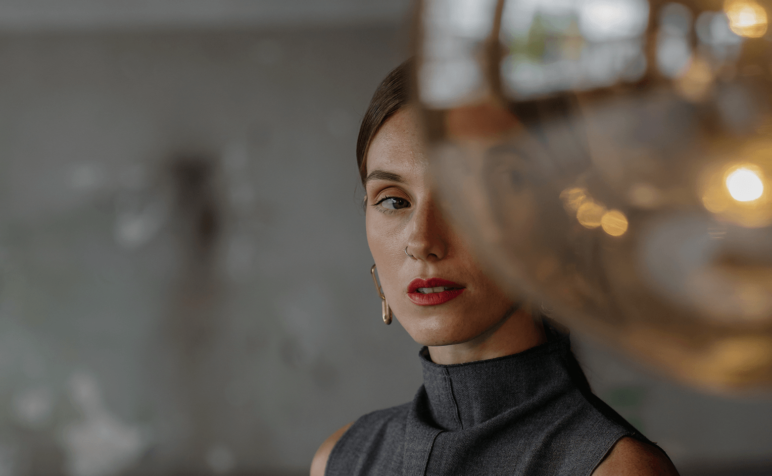 Woman in a sleeveless grey outfit partially obscured by a glowing pendant light, with a soft industrial background