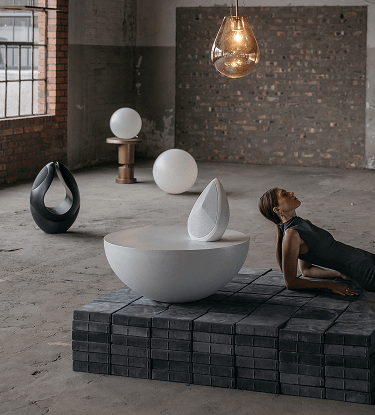 obsidian white wireless speaker on sculptural table beside woman in minimalist industrial space, with Pantheone I black speaker in background