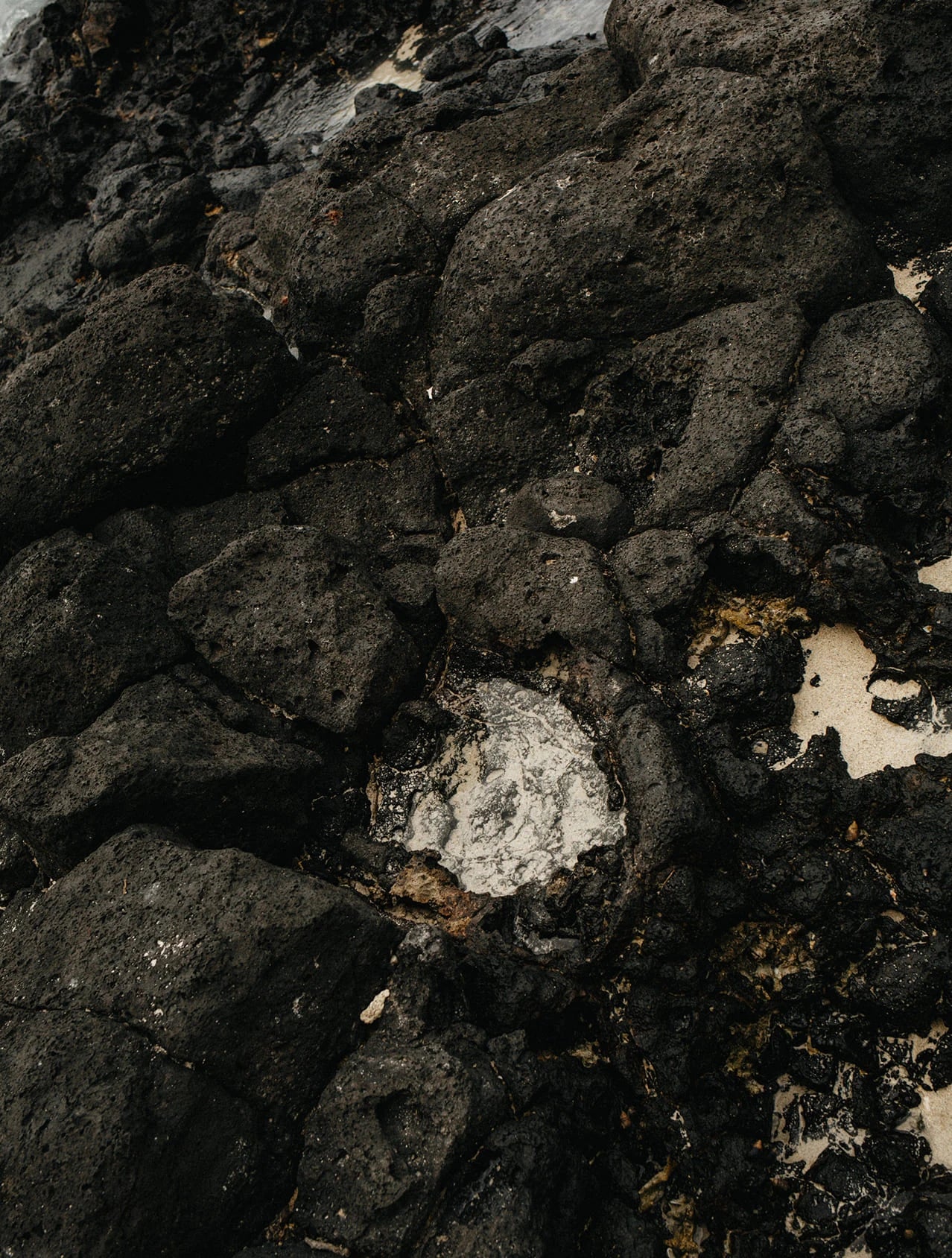Close-up of dark volcanic rock with rough black and charcoal surfaces, some areas showing light gray mineral deposits and small patches of pale sand or foam.