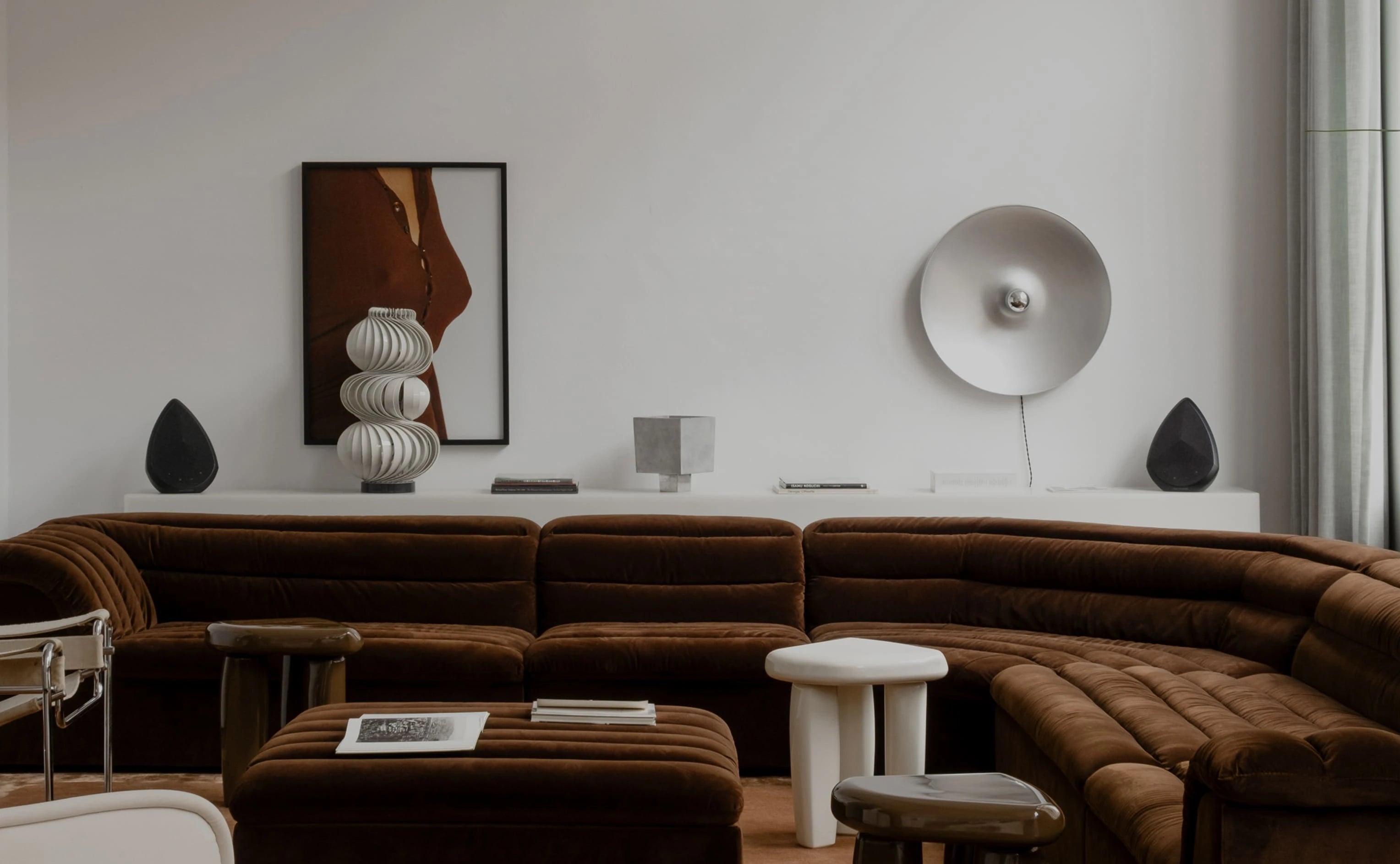A modern living room featuring the Obsidian wireless speakers in a stereo pair mode.With a sofa with matching ottomans, styled with white side tables, sculptural wall lamps, and a framed abstract print on a light grey wall.