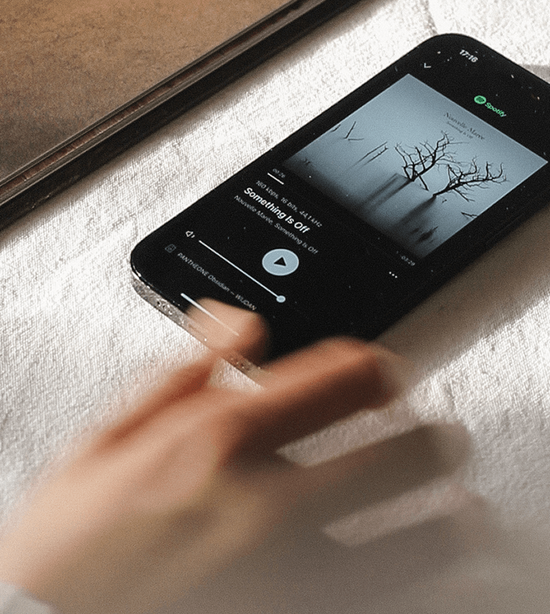 Close-up of a smartphone on a white surface, displaying the Pantheone app playing 'Something Is Off' by Nouvelle Vague. A hand is partially visible in the foreground