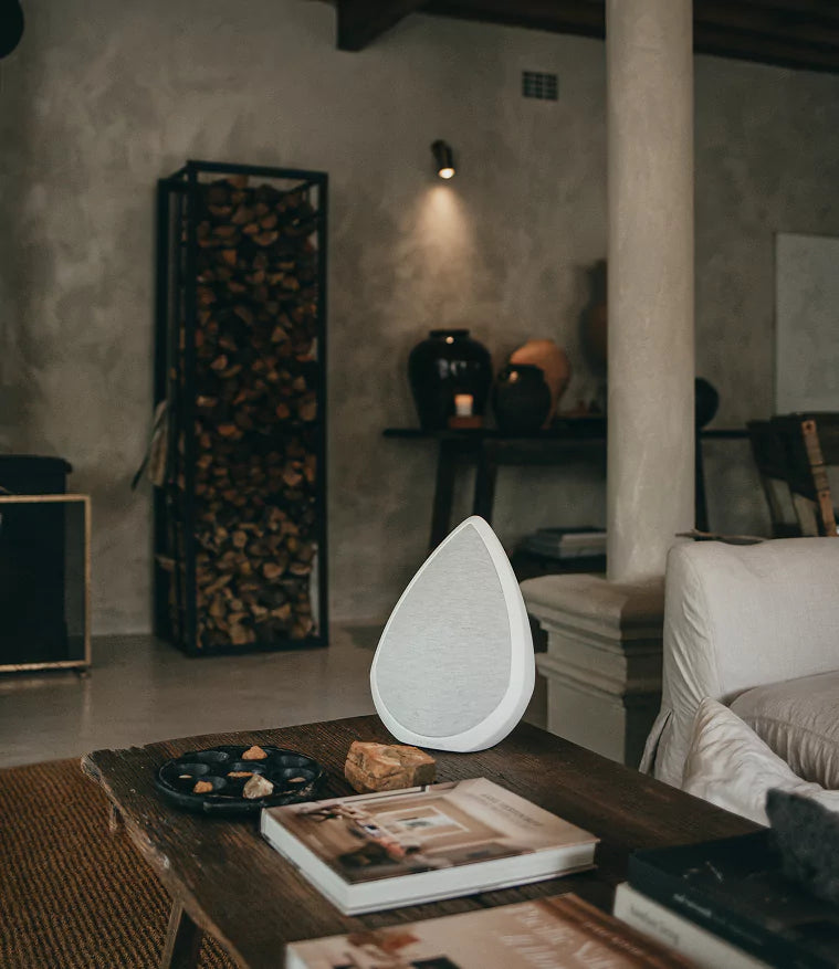 A white, teardrop-shaped Pantheone Obsidian speaker sits on a rustic wooden coffee table in a dimly lit, cozy living room. A stack of books and a small decorative plate with stones are also on the table. In the background, there's a tall, black metal log holder filled with firewood, and a spotlight illuminates a textured wall with decorative vases