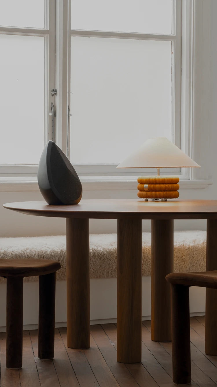 The Obsidian speaker on a wooden table with a lamp and chair in a bright room