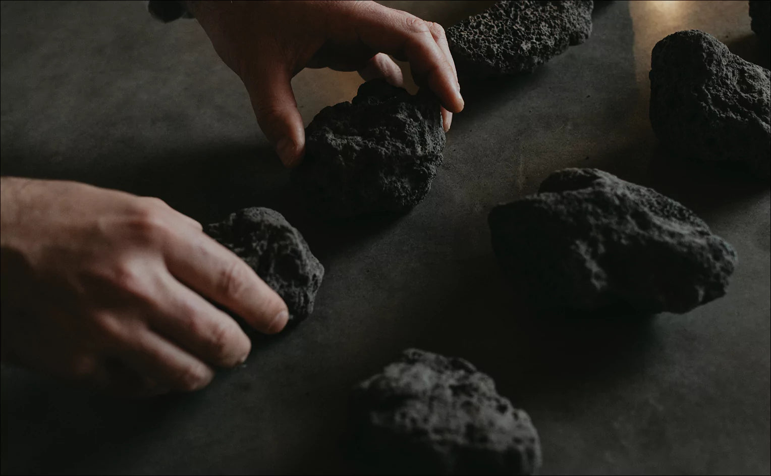 A close-up shot of two hands arranging several rough, dark grey or black porous rocks on a smooth, dark surface. One hand holds a rock while the other is reaching for another, suggesting a thoughtful arrangement or selection of the natural elements