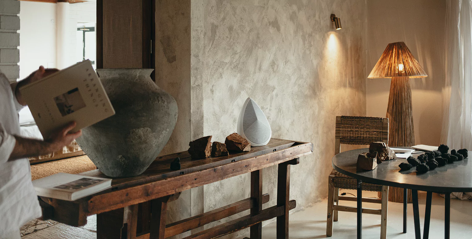 A white,  Pantheone Obsidian portable speaker sits on a rustic wooden console table, alongside a large grey ceramic pot and several natural rock formations. In the foreground, a person holds an open book, partially obscured. The room features textured, light-colored walls, a woven floor lamp, and a round table with wicker chairs, creating an earthy, minimalist aesthetic